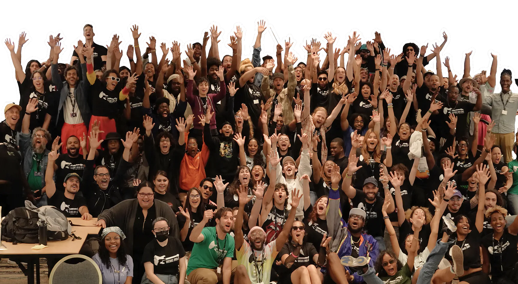 A large group of Guitars Over Guns students, mentors, and staff posing together with their hands raised in celebration against a bright blue background.